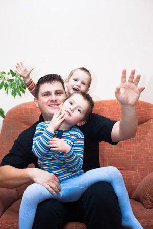 Portrait of happy children boys and daddy waving and sitting on sofa at homeの写真素材