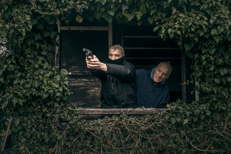 Two masked armed men hiding on overgrown porch of old cabin.の写真素材
