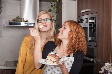 Happy girlfriends enjoying cake and feeding each other in the kitchen at home.の写真素材