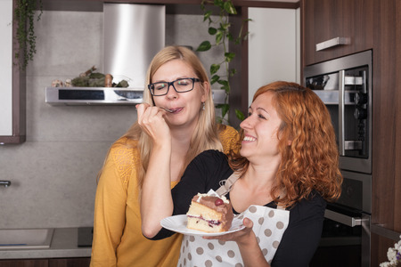 Delighted girlfriends enjoying cake and feeding each other in the kitchen at home.の写真素材