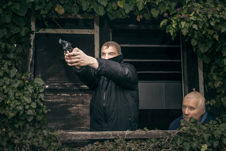 Two masked armed men hiding on overgrown porch of old cabin.の写真素材