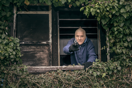 Masked senior escapee man aiming a gun, hiding on overgrown porch of old cabin.の写真素材
