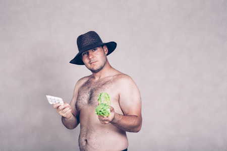 Naked corpulent man in hat holding pharmaceutical products and lettuce.の写真素材