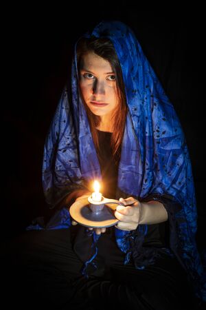 Sad young woman with candle and blue headscarf against black backgroundの写真素材