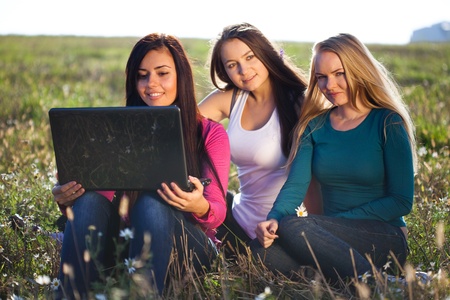 three young beautiful woman with a laptop sitting in the field on sky background の写真素材