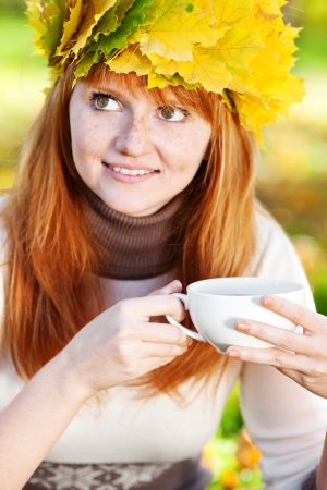 young redhead teenager woman in a wreath of maple leaves with cup of teaの写真素材