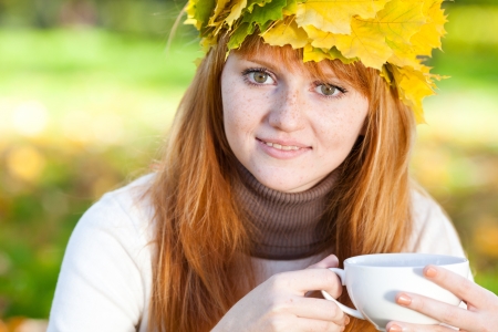 young redhead teenager woman in a wreath of maple leaves with cup of teaの写真素材