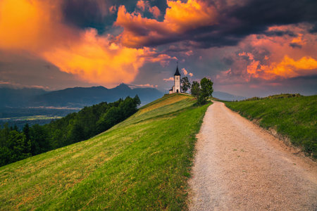 Magical sunset landscape with cute church on the hill. Alpine landscape and gorgeous charming Saint Primoz church with mountains in background at sunset, Jamnik village, Slovenia, Europeの写真素材