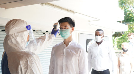 Disease control experts  use an Infrared thermometer equipment to check the temperature on the forehead and use alcohol gel to screen the patients addicted to Covid before entering the building.の写真素材