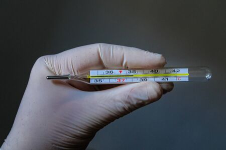 Hand of medic wearing white latex glove holding thermometer which shows high temperature on a dark background. Coronavirus disease 2019 (COVID-19).の写真素材