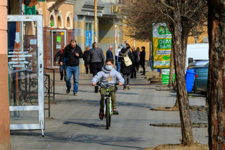 Uzhgorod, Ukraine - March 18, 2020: A boy in a medical mask rides a bicycle in the city center...