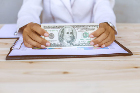 Business woman gripping The United States hundred-dollar bills in her hand and some put on the table full with business charts and notebookの写真素材