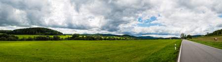 The road to the small ancient city in Czech. The panoramic view of beautiful landscape with pasture, meadow, field, forest, road and church on horizon. Cloudy weather.の写真素材