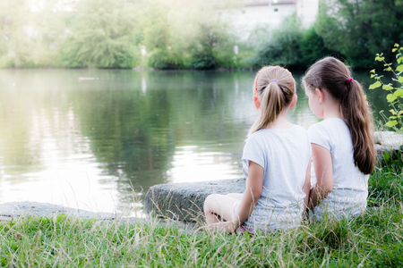 Two beautiful young girls are sitting near pond and looking to the water surface. On background are trees, gras and water surface. Foggy and misty place.の写真素材