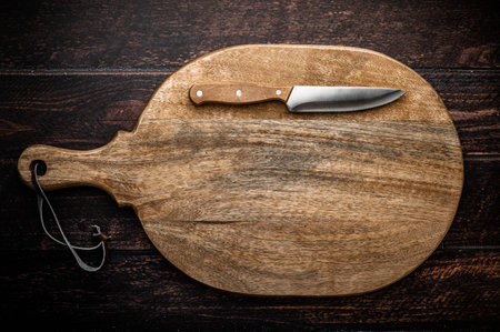 A large wooden cutting board with a knife on a dark wooden table.