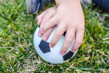 little boy is holding a ball with his both hands on green grass of a playground