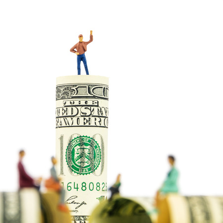 defocused miniature figurines sitting on the edge of 100 dollar banknote with a man standing on dollar bill at the back on white backgroundの写真素材