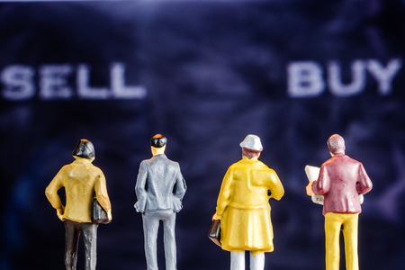 closeup of miniature figurines of businessmen standing in front of big defocused screen with sell and buy words forming a backgound patternの写真素材