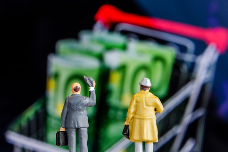 closeup of miniature figurines of businessmen standing in front of big defocused screen with 100 euro banknotes in shopping rolleyの写真素材