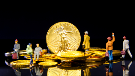 closeup of golden american eagle coin on black background with many golden coins at bottom and miniature figurines standing aroundの写真素材