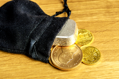 closeup of golden coins and silver brick falling out of black moneybag and laying on wooden backgroundの写真素材