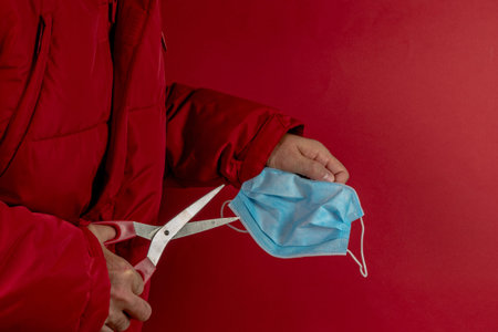 man in red jacket trying to cut a protective mask in half against corona virus on red background,...