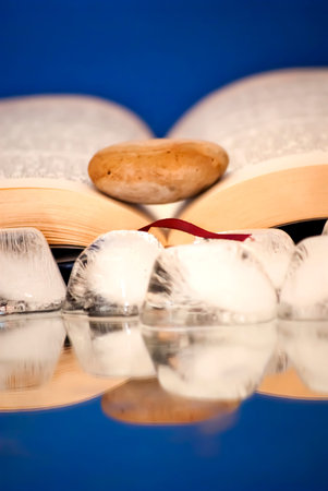 Book open with a single stone on it over a mirror with ice cubes with blue background