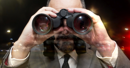 Handsome business man, bald and bearad, looking through binoculars through a window into the city at nightの写真素材