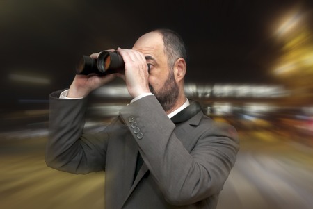 Handsome business man, bald and bearad, looking through binoculars over 
a background of an office building lit in the nightの写真素材
