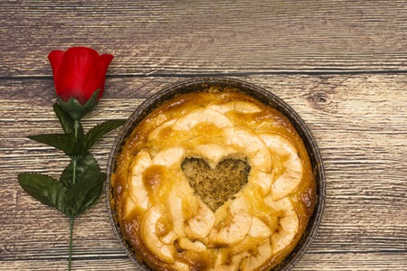Apple pie with a hollow in the shape of a heart on a wooden table in which there is a red roseの写真素材