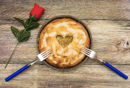 Apple pie with a hollow in the shape of a heart and two forks on a wooden table in which there is a red roseの写真素材