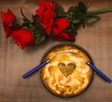 Apple pie with a hollow in the shape of a heart and two forks interlaced on a wooden table in which there is a bouquet of red rosesの写真素材