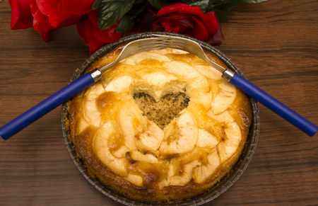 Apple pie with a hollow in the shape of a heart and two forks interlaced on a wooden table in which there is a bouquet of red rosesの写真素材