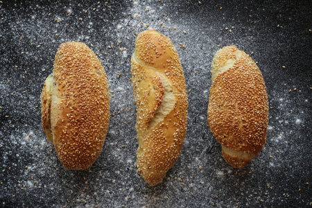 Flat lay shot of three fresh corn bread rolls with sesame seeds on black sprinkled with flour and...