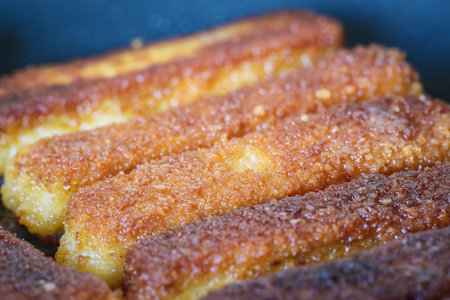 Fish fingers fry on frying pan with golden crunchy coating, closeup with narrow depth of field