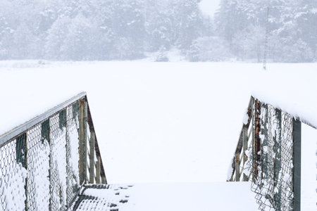 Metal footbridge over small river covered in snow during snowfall lead to field