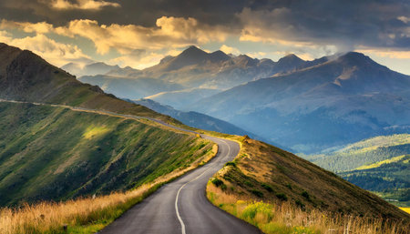 A winding road meanders through rolling hills under a dramatic sky, leading towards towering mountain peaks in the background, creating a breathtaking natural landscape.の写真素材