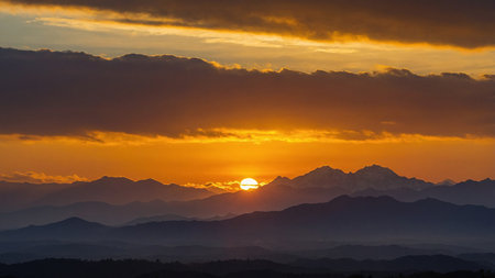 The image captures a breathtaking sunset over rolling mountain ranges, with mist enveloping the valley and a sky bathed in golden hues, evoking a sense of tranquility and grandeur. AI generated imageの写真素材