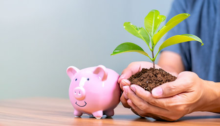 Hands holding piggy bank with green plant growing out of soilの素材