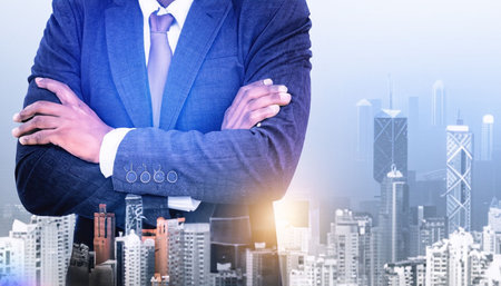Double exposure of confident businessman with arms crossed over modern cityscape background.の素材