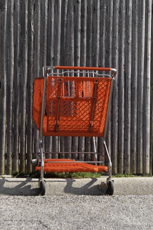A red shopping cart that has been abandoned on a far sdie of a parking lot and near a wooden fence. のeditorial素材