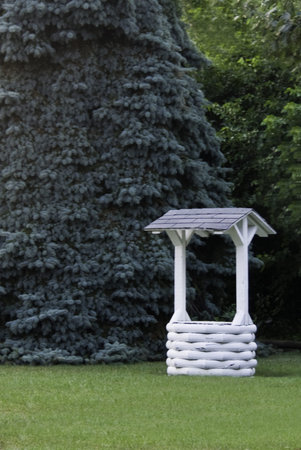 A white painted wishing well in front of a giant evergreen tree. 