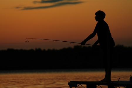 Silhouette of a boy against sunset as he fishes on the pier.の写真素材