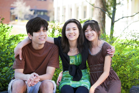 Three biracial brother and sisters sitting outdoors on log, smiling