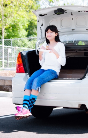 Preteen biracial girl sitting in opened car trunk, eating lunchの写真素材