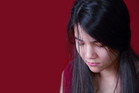 Beautiful, biracial teen girl looking down, depressed or sad, on red backgroundの写真素材