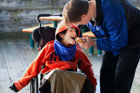 Father feeding disabled son a hamburger in wheelchair. Child has cerebral palsyの写真素材