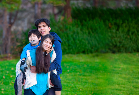 Disabled biracial child riding piggy back on his sister, family surrounding him, together at park.の写真素材