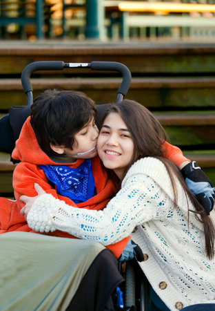 Disabled little boy kissing his big sister on cheek while seated in wheelchairの写真素材
