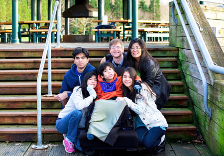 Interracial family surrounding disabled boy in wheelchair outdoors by stairs, child has cerebral palsyの写真素材